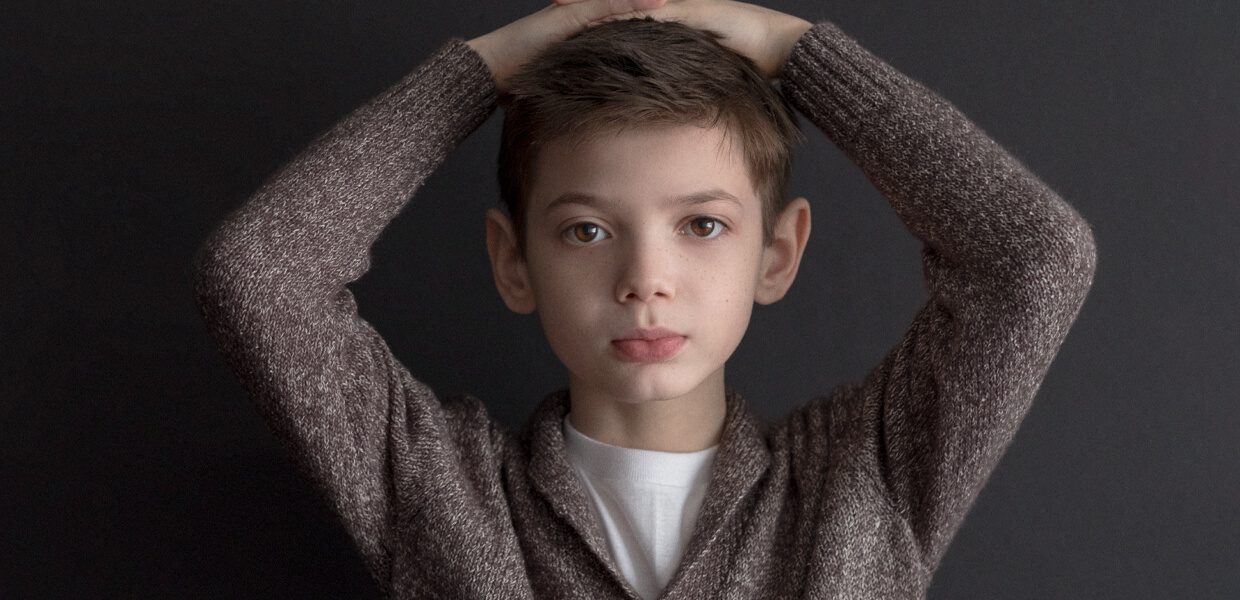 Portrait of a young boy with hands behind his head, wearing a brown sweater.