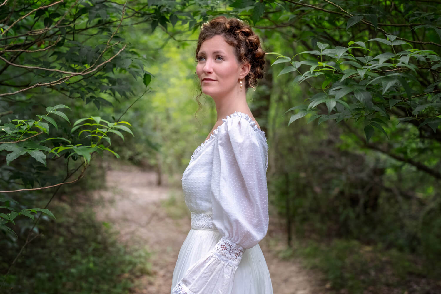 Woman in a white vintage dress standing in a lush green forest path.